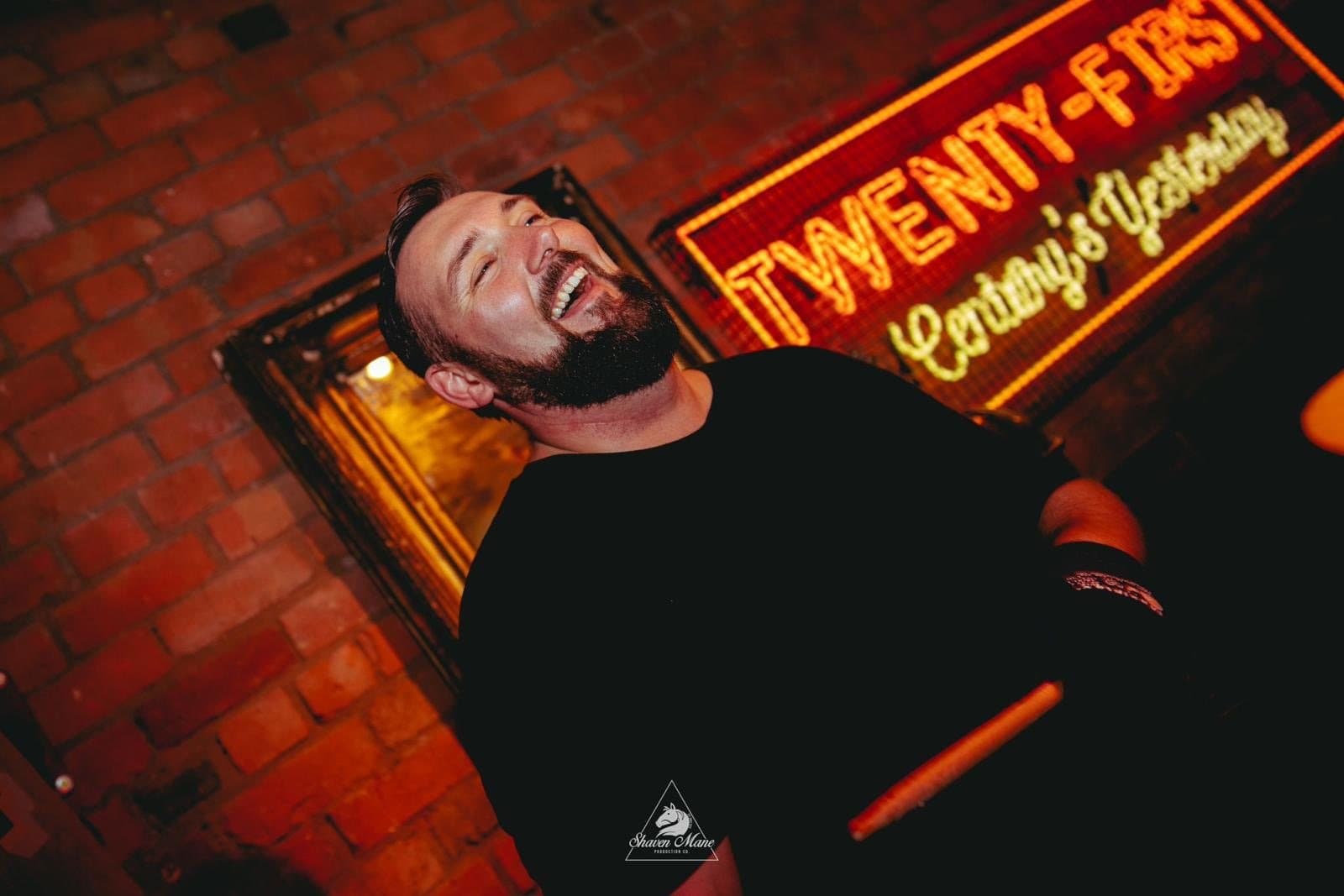 A drummer laughing at the kit under a neon sign.
