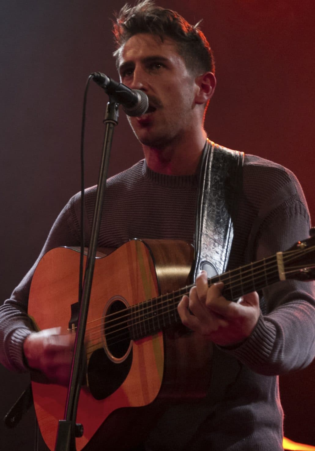 Jamie Duff singing into a mic and playing acoustic guitar under red stage light.