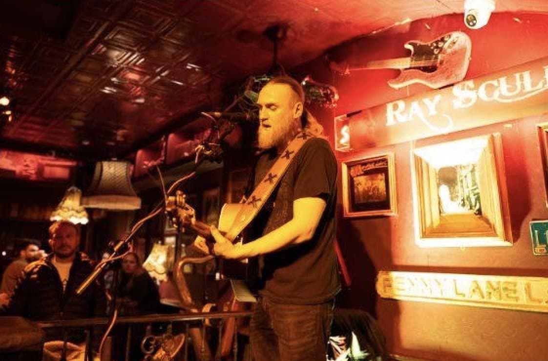 An acoustic guitarist playing at floor level with drinkers close by.